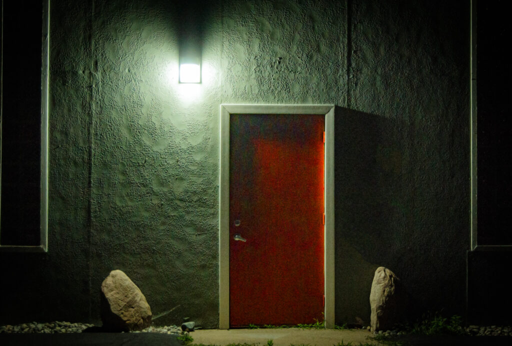 A liminal space showing a vibrant red door illuminated by a cool white porchlight. The door is set into a brick wall with a large rock on both sides.