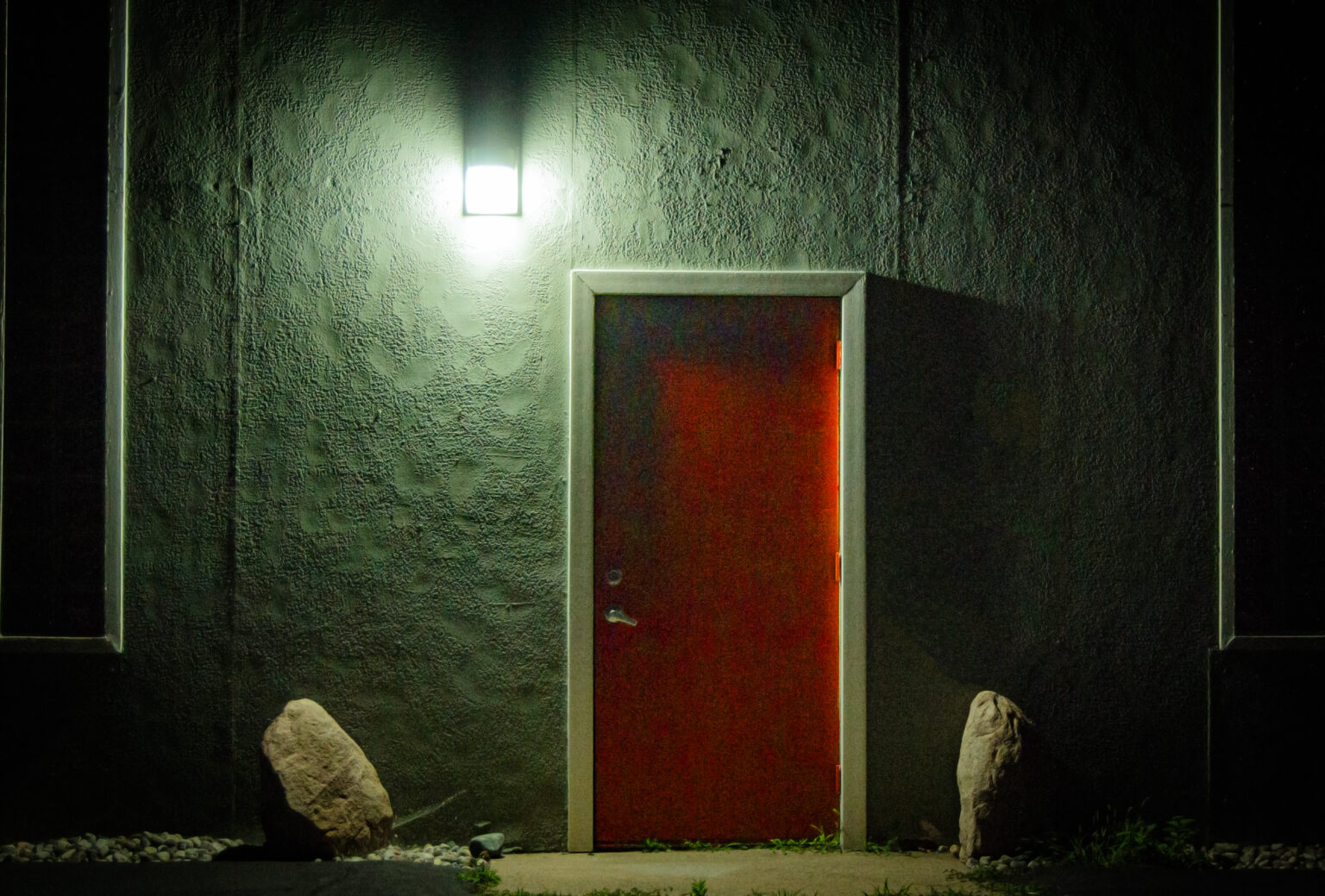 A liminal space showing a vibrant red door illuminated by a cool white porchlight. The door is set into a brick wall with a large rock on both sides.